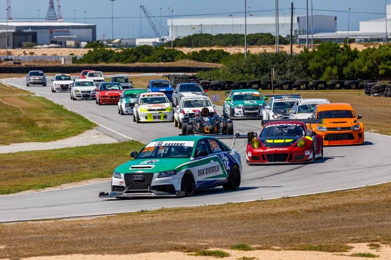 Large mixed-class field of touring and sports cars rounds a corner at Aldo Scribante Raceway with Port Elizabeth harbor in the background