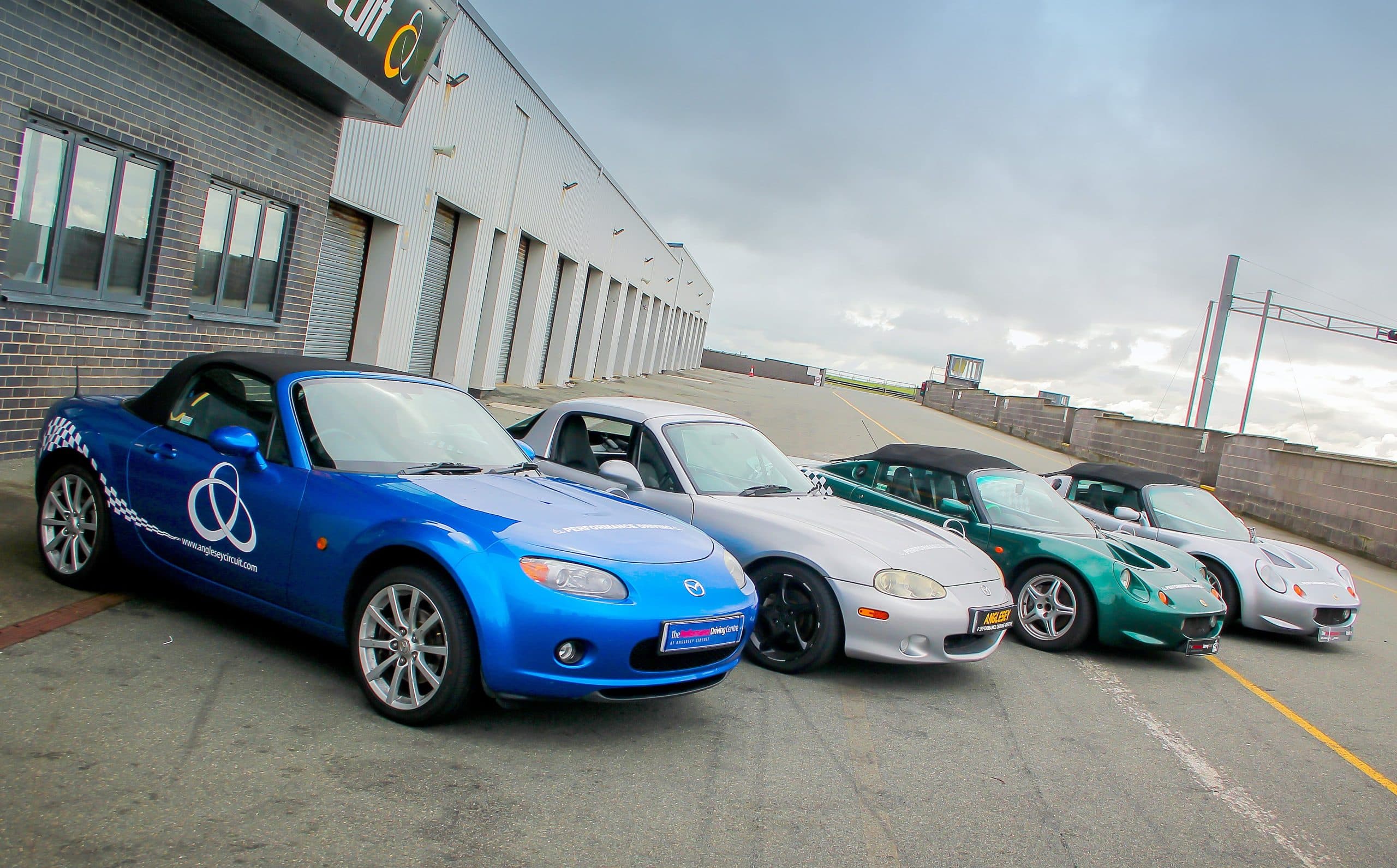 Three sports cars including a blue Mazda MX-5 and Lotus lined up outside the pit building at Anglesey Circuit, Wales