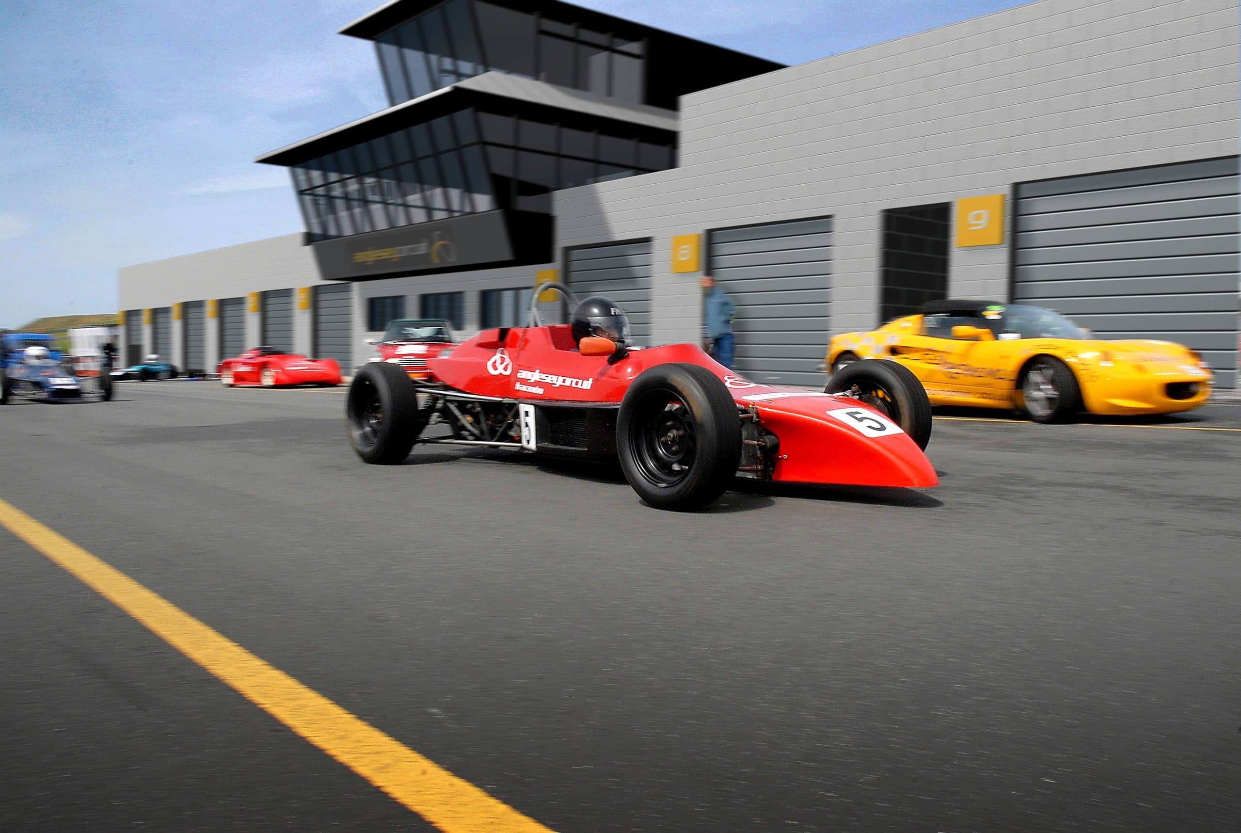 Red Formula Ford single-seater in the pit lane at Anglesey Circuit, with the modern pit building and control tower behind