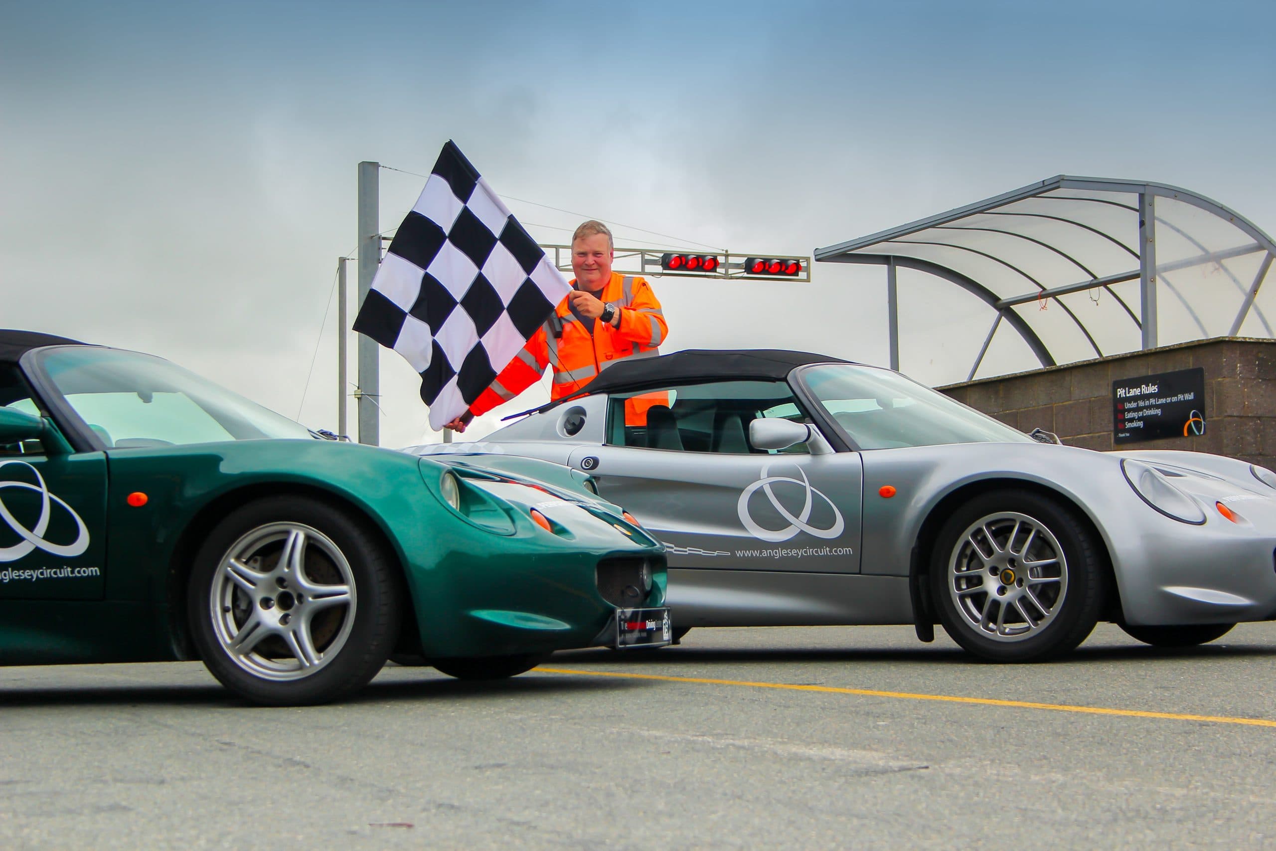 Anglesey Circuit marshal waves a chequered flag beside two Lotus Elise sports cars in the pit lane at Trac Mon, Wales