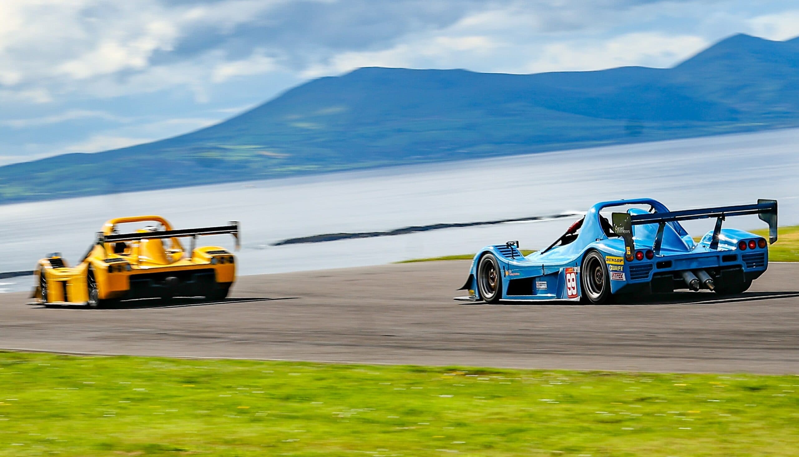Blue and yellow sports prototype cars racing at speed at Anglesey Circuit with sea and Snowdonia mountains in the background