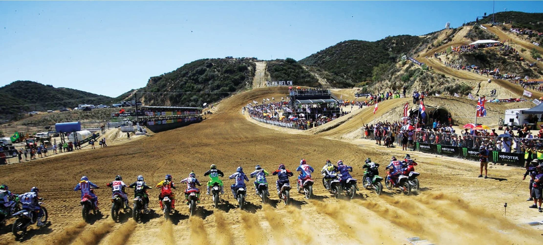 Full gate of motocross riders charging into the first turn at Glen Helen Raceway during a Monster Energy national event, packed hillside grandstands visible