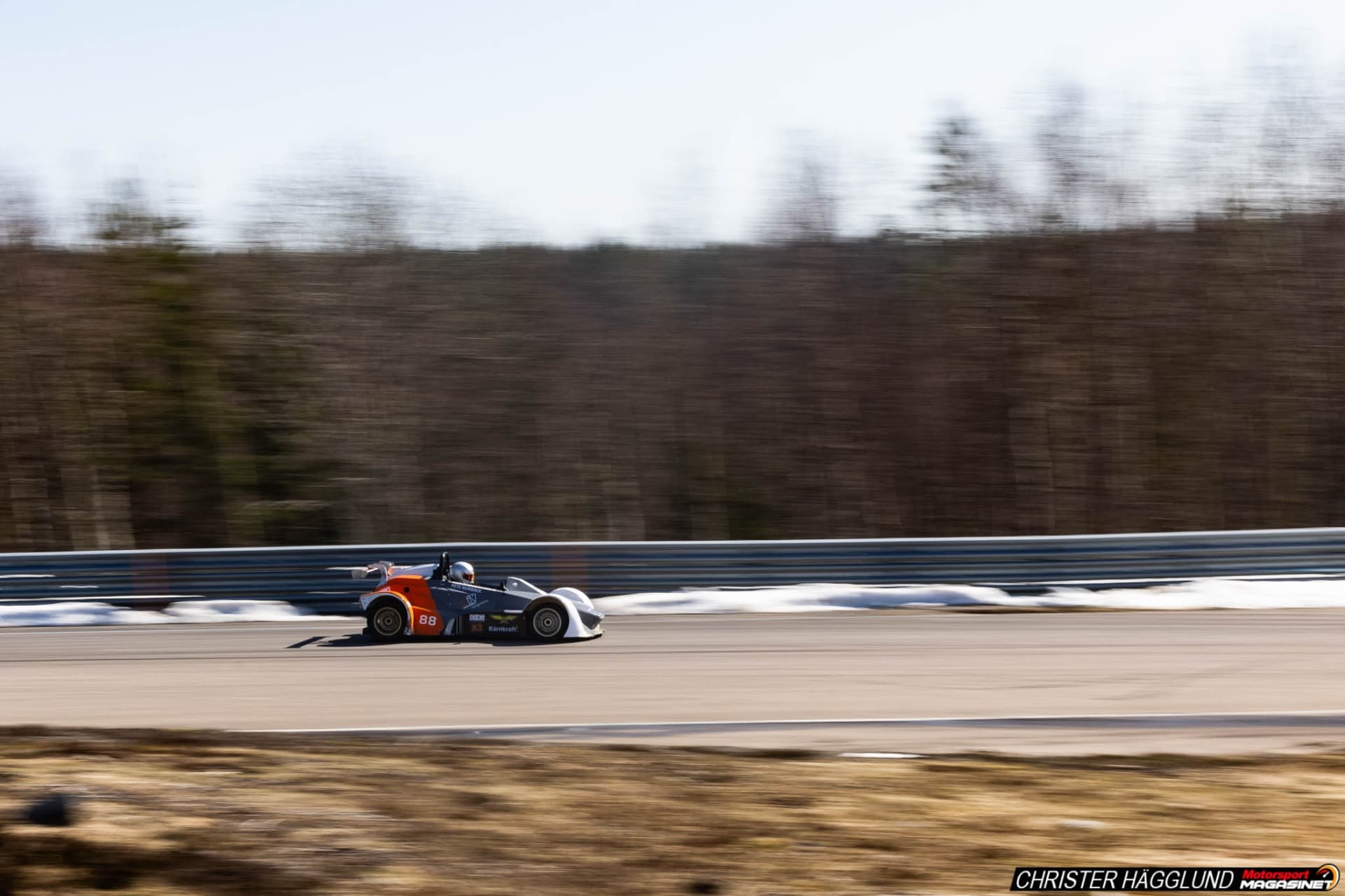 Orange prototype race car at speed on the Mittsverigebanan circuit near Härnösand, Sweden, pine forest in background