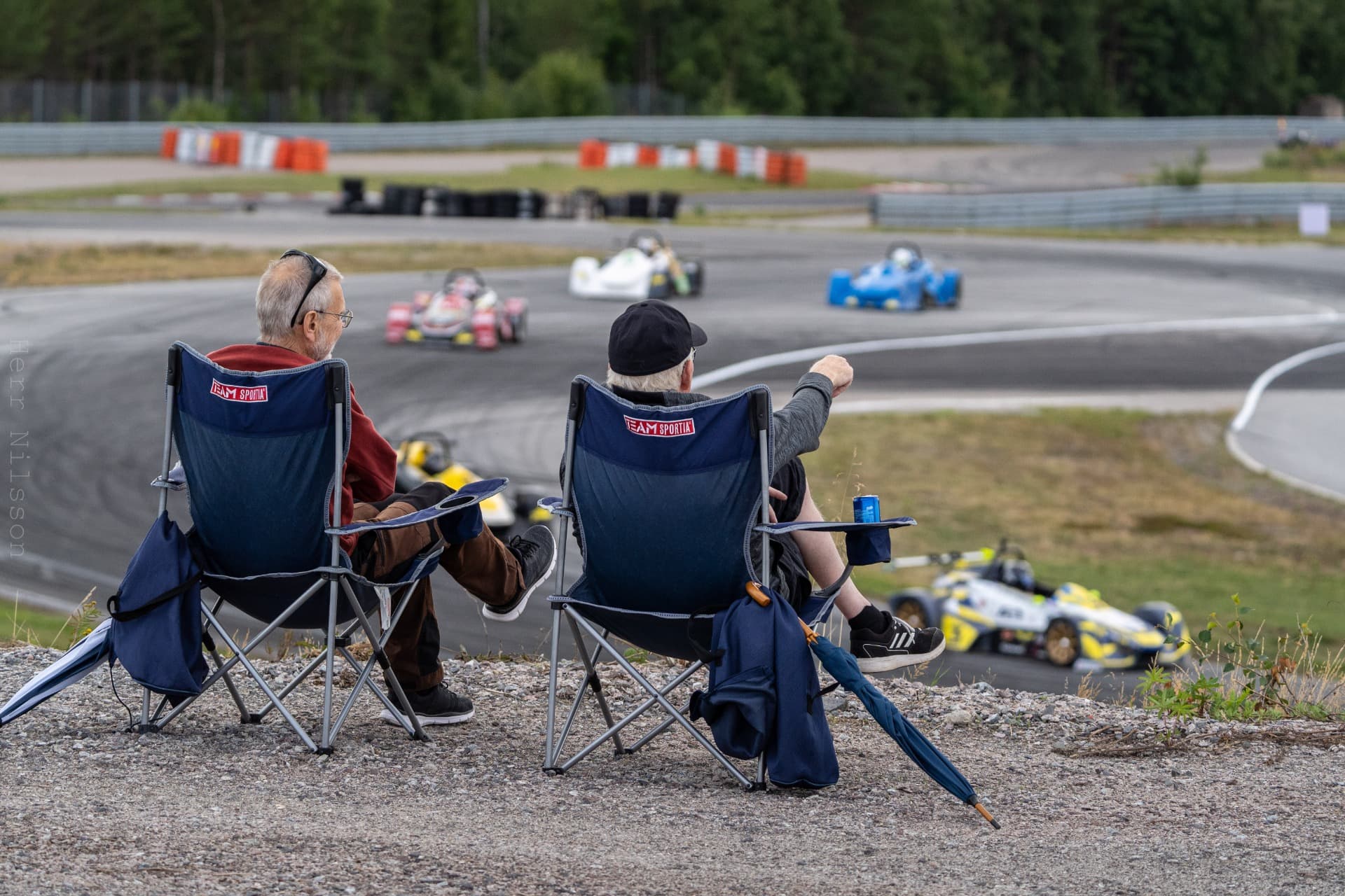 Two spectators in folding chairs watching formula cars race through a corner at Mittsverigebanan, Härnösand, Sweden