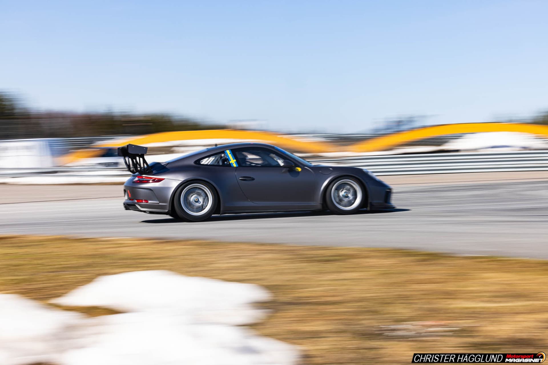 Dark gray Porsche 911 GT3 at speed along the barrier wall on Mittsverigebanan in early spring, Härnösand, Sweden