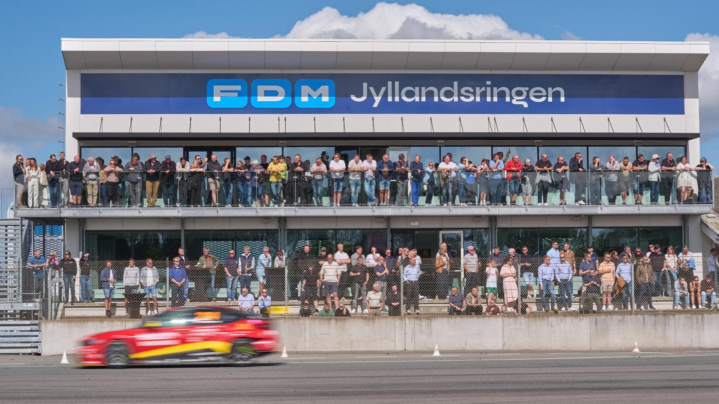 Spectators lining the FDM Jyllandsringen grandstand terrace as a red race car blurs past below